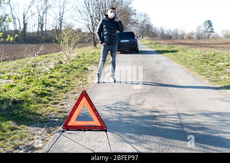 triangolo segnaletico rosso sulla strada davanti a un'auto rotta. Un giovane che usa un telefono per ordinare il servizio di assistenza Foto Stock