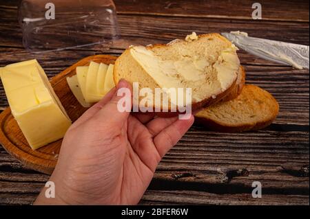 Qualcuno spalma il burro su pane tostato di grano fresco con un coltello e un piatto di burro di legno con un pezzo di burro e fette di formaggio su uno sfondo di legno. Foto Stock