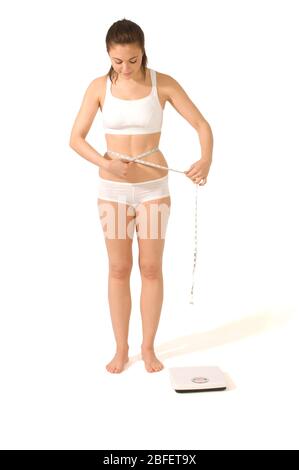 Una giovane donna che misura lo stomaco con un nastro di misurazione, mentre in piedi accanto ad una bilancia da bagno - modello rilasciato Foto Stock
