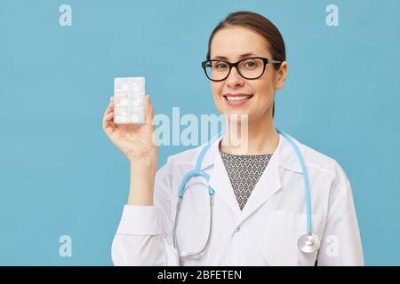 Ritratto di medico femminile in occhiali tenendo pillole in mano e sorridendo alla macchina fotografica isolato su sfondo blu Foto Stock