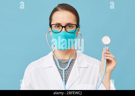Ritratto di medico femminile in maschera protettiva che tiene stetoscopio e guardando la fotocamera su sfondo blu Foto Stock