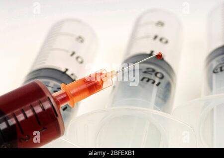 Vista di un ago fissato ad una siringa, la punta dell'ago contiene una goccia di sangue, di fronte ad uno sfondo di siringhe monouso non utilizzate. (Io Foto Stock