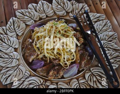 Carne di coniglio cotta con pasta, cipolle e altre spezie su un tavolo di bambù. Pinze per pasta e forchetta pronti. Foto Stock