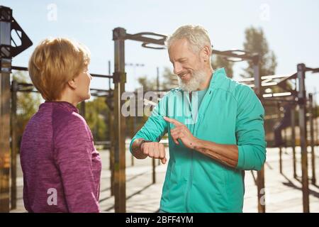 Verifica dei risultati del fitness. Buon uomo senior in abbigliamento sportivo guardando lo smartwatch e sorridendo mentre si esercita con sua moglie in palestra all'aperto. Attivo Foto Stock