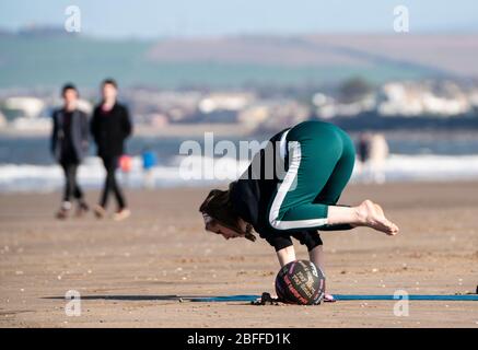 Portobello, Scozia, Regno Unito. 18 aprile 2020. Vista sul lungomare di Portobello e sulla spiaggia su un sole ma freddo e ventoso Sabato pomeriggio durante il blocco dei coronavirus nel Regno Unito. Donna pratica yoga sulla spiaggia. Iain Masterton/Alamy Live News Foto Stock