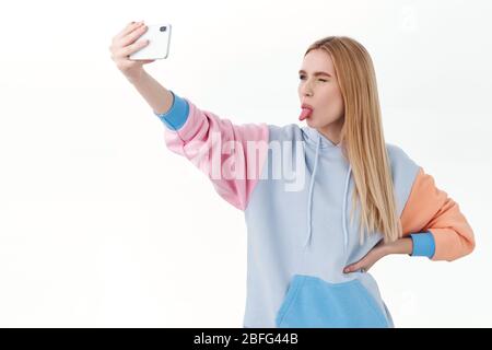 Comunicazione, tecnologia e concetto online. Sciocco attraente vlogger femminile, ragazza carina con cappuccio, prendendo selfie su smartphone con braccio esteso Foto Stock