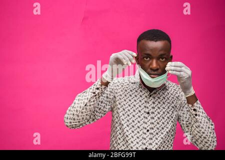 un giovane medico africano isolato su sfondo rosa cercando di indossare maschera facciale per prevenire, prevenire, si è impedito dallo scoppio in questo modo Foto Stock