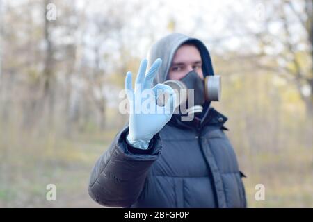 Ritratto di giovane uomo in maschera protettiva gas e guanti monouso in gomma mostra un gesto okay all'aperto in legno di primavera. Concetto di prodotti di protezione usag Foto Stock