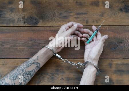 Mani maschili in manette e siringa con dose di farmaco su tavolo di legno. Concetto di persona tossicodipendente arrestato Foto Stock