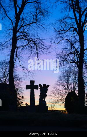 Silhouette di un angelo e croce di testa con gli alberi in un cimitero all'alba. Kings Sutton, Northamptonshire, Inghilterra. Silhouette Foto Stock