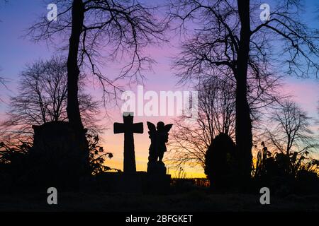 Silhouette di un angelo e croce di testa con gli alberi in un cimitero all'alba. Kings Sutton, Northamptonshire, Inghilterra. Silhouette Foto Stock
