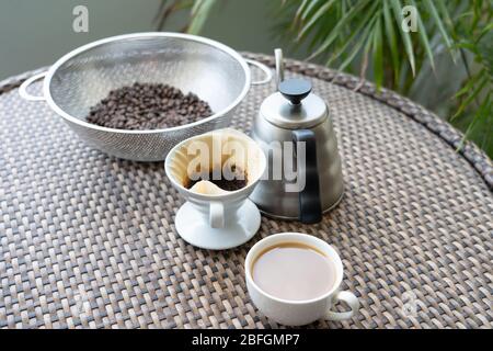 Una tazza di caffè sul tavolo con chicchi di caffè appena tostati, filtro antigoccia e bollitore Foto Stock