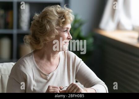 Donna anziana sorridente seduta sul divano che guarda fuori dalla finestra Foto Stock