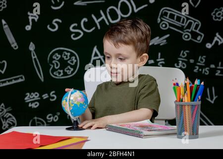 Carino ragazzo intelligente è seduto a una scrivania con globo in mano sullo sfondo con lavagna . Pronto per la scuola. Ritorno a scuola Foto Stock