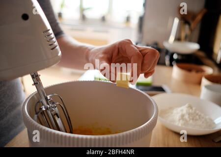 Giovane donna che cuoce una torta di gemme in cucina in piedi al bancone utilizzando un mixer portatile per frullare gli ingredienti più freschi in una ciotola bianca. Foto Stock