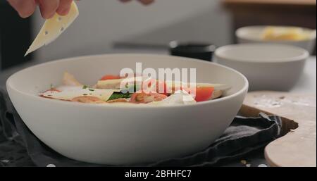 man hand aggiunge fiocchi di formaggio su insalata con pollo, romaina e pomodori in ciotola bianca Foto Stock