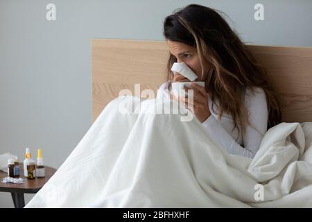 Giovane donna malata che soffia il naso, tenendo il fazzoletto, seduta a letto Foto Stock