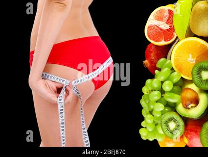 Bella giovane donna che misura il suo corpo con nastro e frutti tropicali vicino a lei isolato su nero Foto Stock