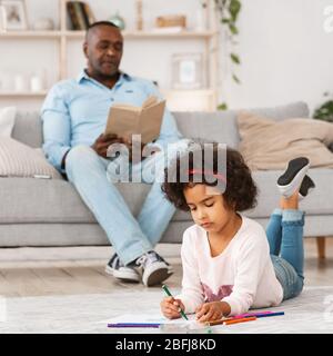 Momenti di tranquillità a casa. Bella ragazza che disegnava sul pavimento e suo nonno che legge interessante libro in soggiorno Foto Stock