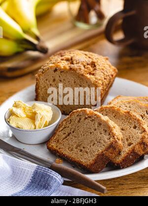 Torta di pane di banana-noci tagliata e burro su un piatto con un mazzo di banane in background Foto Stock