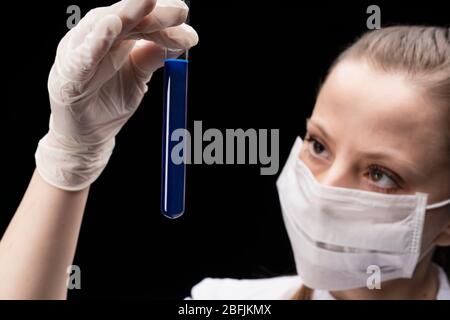 una provetta con una sostanza luminosa blu liquida nelle mani di una giovane donna in una maschera, uno specialista scientifico o studente laureato Foto Stock