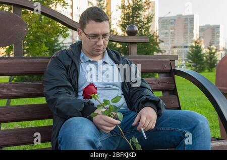 un ragazzo triste, accovigante, sconvolto con una sigaretta fumante e una rosa rossa in mano si siede su una panchina in un parco cittadino Foto Stock