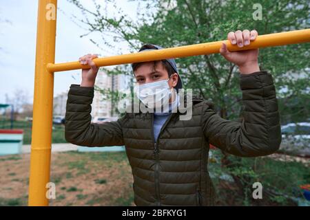 teen boy si trova vicino al bar orizzontale di attrezzature per l'allenamento all'aperto, sul parco giochi vicino a edifici alti con appartamenti, una maschera medica sul suo viso p Foto Stock