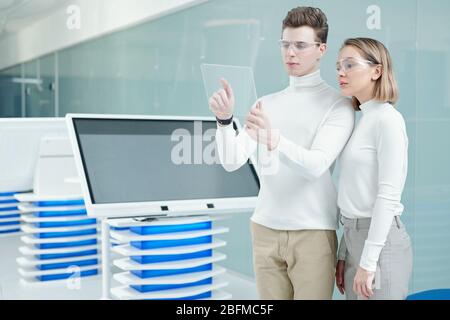 Giovane uomo intelligente e sua collega con occhiali intelligenti che guardano il display di un tablet trasparente mentre scansionano nuove informazioni Foto Stock