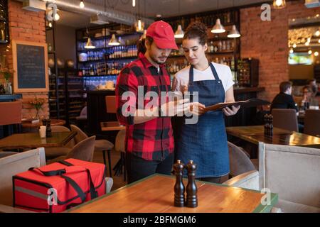 Corriere giovane con smartphone e cameriera con menu in piedi al tavolo e controllo degli ordini online prima di consegnarli ai clienti Foto Stock