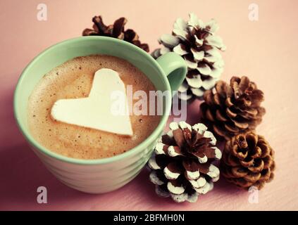 Tazza di cappuccino caldo con marshmallow al cuore, mitrini caldi e coni di pino su sfondo rosa Foto Stock