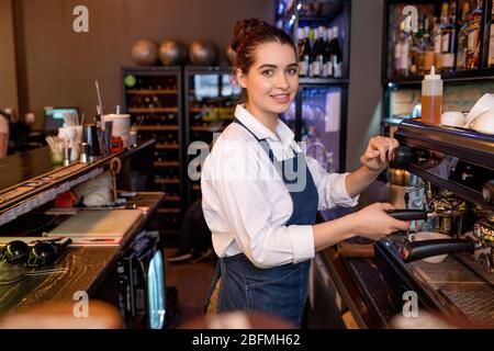 Cameriera gentile e giovane in abbigliamento da lavoro che prepara cappuccino mentre si trova accanto alla macchina da caffè nella caffetteria o nel ristorante Foto Stock