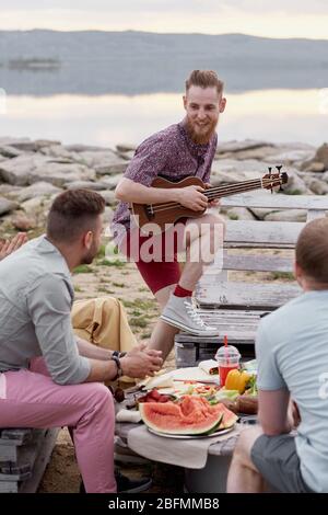 Giovane uomo bearded che suona la chitarra e sorridente felicemente mentre trascorre la serata estiva con gli amici all'aperto vicino al lago Foto Stock