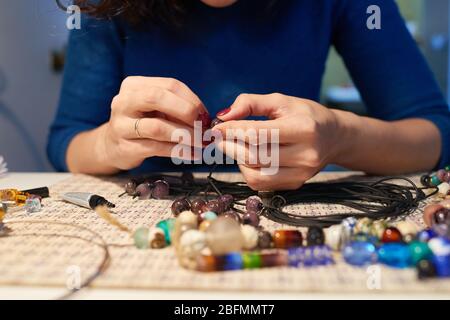 Mani di giovane donna creativa seduta dal posto di lavoro e mettendo le perle di vetro sul filo di plastica mentre fanno le collane in officina Foto Stock