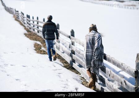 Vista posteriore della giovane donna avvolta in plaid che si muove lungo recinto mentre segue il suo ragazzo la mattina invernale fuori Foto Stock
