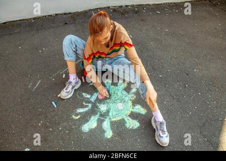 La ragazza teenage siede su uno skateboard e disegna il coronavirus con gesso sul marciapiede Foto Stock