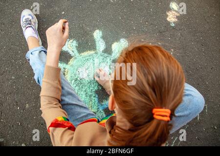 La ragazza teenage siede su uno skateboard e disegna il coronavirus con gesso sul marciapiede Foto Stock