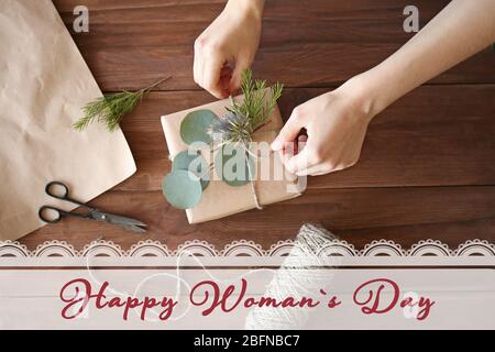 Mani femminili che fanno la scatola del regalo con i fiori sul tavolo di legno. Testo GIORNO DELLA DONNA FELICE sullo sfondo Foto Stock