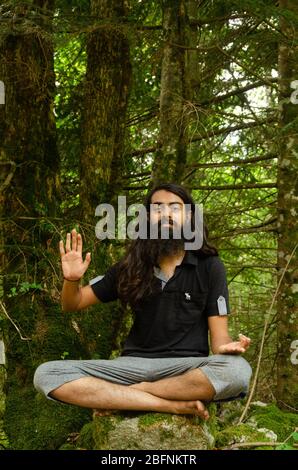 Bearded e capelli lunghi indiano ragazzo in posa blessing seduta croce gambe su una roccia contro la natura foresta sfondo Foto Stock