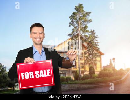 Agente immobiliare in possesso di un cartello con testo IN VENDITA e chiave di fronte a casa di campagna all'aperto Foto Stock