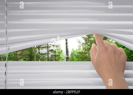 Donna che separa le stecche di ciechi e guardando attraverso la finestra a bellissimo paesaggio Foto Stock