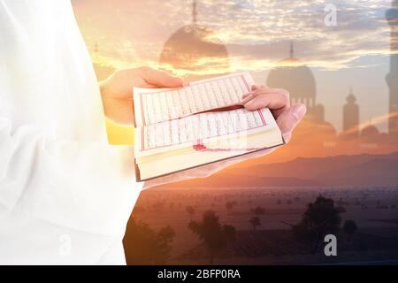 Giovane musulmano che legge Corano e silhouette della moschea con paesaggio sullo sfondo Foto Stock