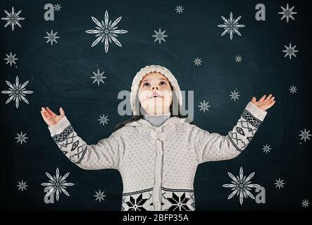 Bambina in un abito lavorato a maglia alla moda guardando in su e disegnato fiocchi di neve su sfondo lavagna Foto Stock