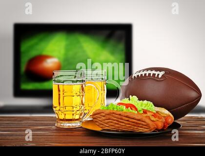 Due bicchieri di birra, palla e spuntino sul tavolo di legno davanti allo spettacolo televisivo di calcio. Guardare la partita di calcio a casa. Foto Stock