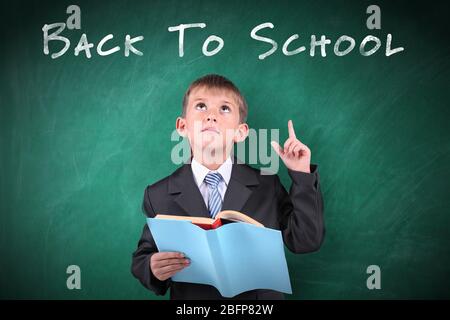 Concetto scolastico. Ragazzo carino con libro in piedi su sfondo lavagna. Testo di ritorno a scuola. Foto Stock