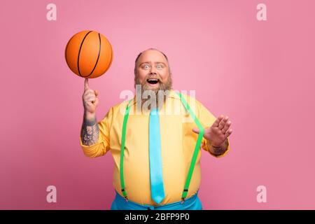 Ritratto del suo simpatico allegro ragazzo bearded girando palla arancione sul dito avendo divertimento isolato su sfondo colore rosa pastello Foto Stock