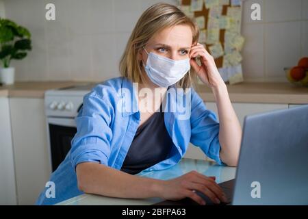 Donna in maschera medica è lavoro ufficio lavoro a distanza da casa in cucina. Tramite computer. Formazione a distanza online e lavoro. Foto Stock