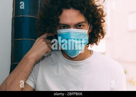 Un uomo con una T-shirt bianca in una maschera medica vuole togliere. È in quarantena domestica durante la pandemia del coronavirus. Concetto di soggiorno a casa. Nuovo vero Foto Stock