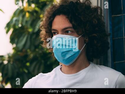 Ritratto di un giovane con capelli ricci lunghi in maschera medica blu e maglietta bianca. Sfondo verde e tenda blu vicino alla finestra. Quarant domestico Foto Stock