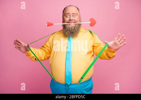 Ritratto del suo simpatico simpatico funky felice ragazzo bearded positivo tenendo in denti bocca freccia amorosa tirando sospendenti avendo divertimento isolato su rosa Foto Stock