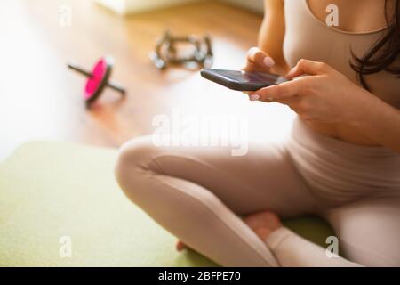 Giovane donna che fa l'allenamento di yoga in camera durante la quarantena. Tagliare la vista della ragazza utilizzando lo smartphone. Donna seduta sul tappetino durante il riposo. Foto Stock